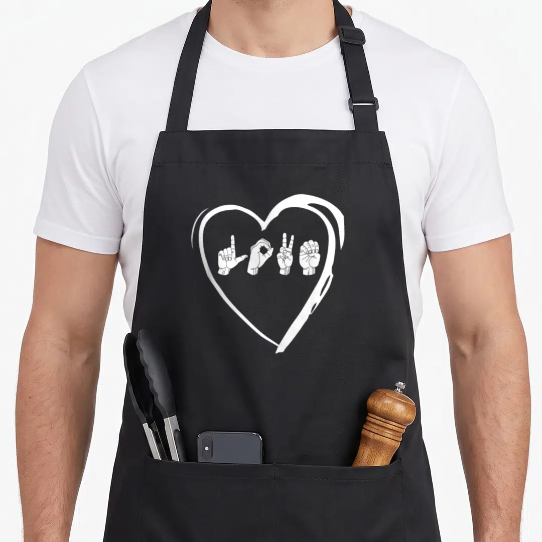 Sign valentine's day apron - White heart outline containing four hands performing ASL signs for L-O-V-E in black line — shown on a black cotton canvas apron against a white background