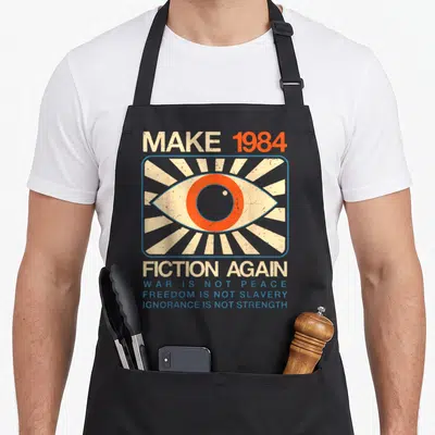 Dystopian fiction apron - Distressed eye graphic with Make 1984 Fiction Again text and blue slogans on black apron — shown on a black cotton canvas apron against a white background