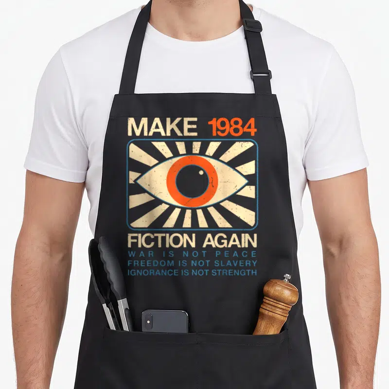 Dystopian fiction apron - Distressed eye graphic with Make 1984 Fiction Again text and blue slogans on black apron — shown on a black cotton canvas apron against a white background