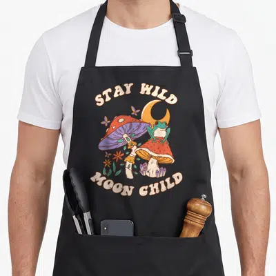 Wild mushroom apron - Frog on red mushroom with crystals, moon, and butterflies under STAY WILD MOON CHILD text — shown on a black cotton canvas apron against a white background