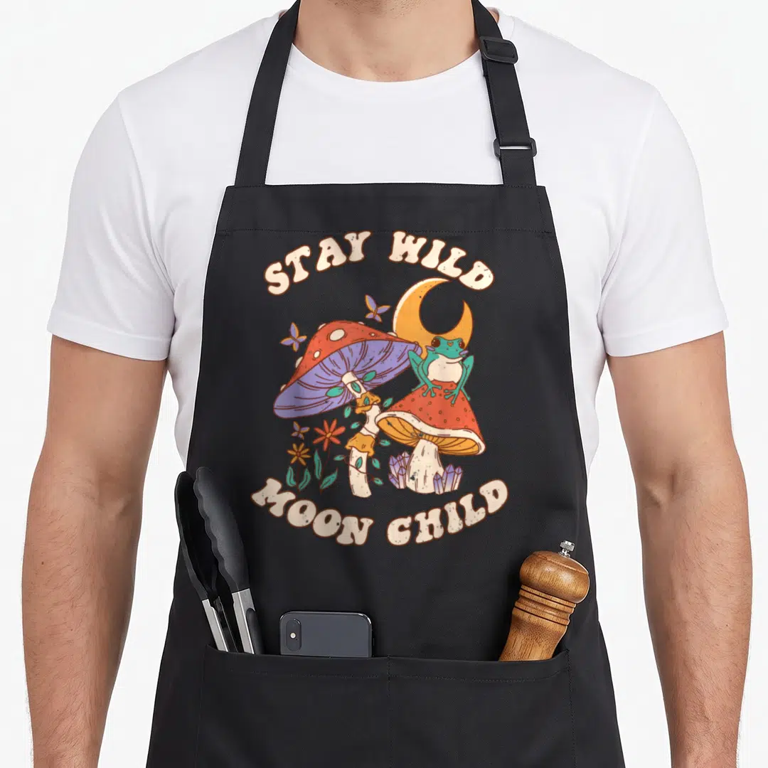 Wild mushroom apron - Frog on red mushroom with crystals, moon, and butterflies under STAY WILD MOON CHILD text — shown on a black cotton canvas apron against a white background