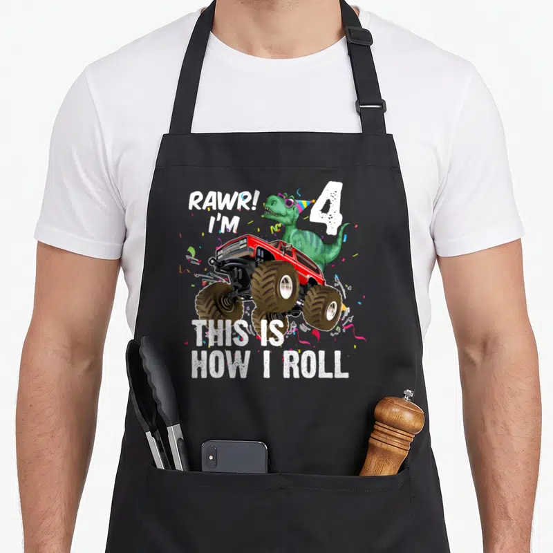 Dinosaur riding truck apron - Green dinosaur in sunglasses driving red monster truck with text Rawr I am 4 This is how — shown on a black cotton canvas apron against a white background