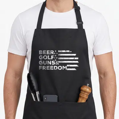 Beer golf apron - White distressed text BEER GOLF GUNS FREEDOM with flag stripes — shown on a black cotton canvas apron against a white background