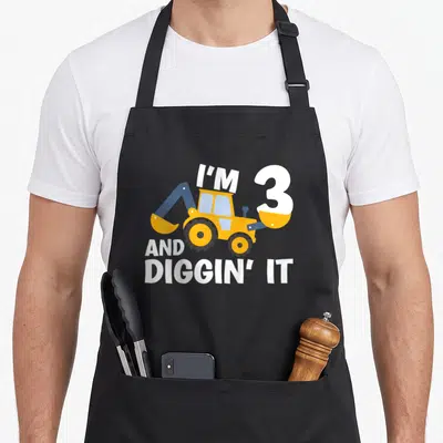 Digging construction apron - Yellow excavator with blue arm and white text reading I'M 3 AND DIGGIN' IT — shown on a black cotton canvas apron against a white background