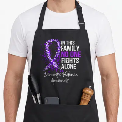 Fight domestic aprons - Purple heart ribbon and In This Family No One Fights Alone text — shown on a black cotton canvas apron against a white background