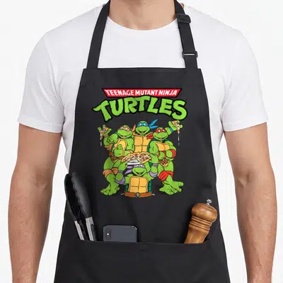 Crew pizza apron - Four cartoon turtles holding pizza under green TURTLES text with red banner — shown on a black cotton canvas apron against a white background