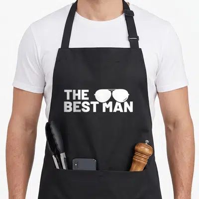 Bachelor supply funny wedding aprons - White THE BEST MAN text stacked with sunglasses overlapping the letters — shown on a black cotton canvas apron against a white background