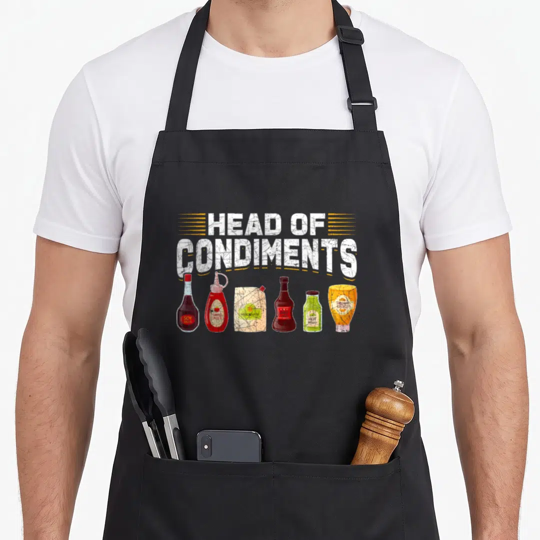 Condiment barbecue apron - Head of Condiments text above six sauce bottles in distressed white, yellow, red, brown — shown on a black cotton canvas apron against a white background