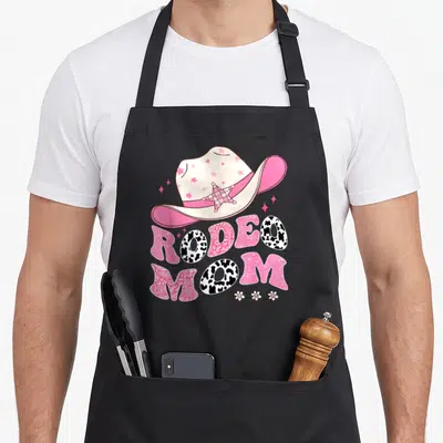 Family cowgirl aprons - Pink cowboy hat above Rodeo Mom text in cow print and paisley patterns with three flowers — shown on a black cotton canvas apron against a white background