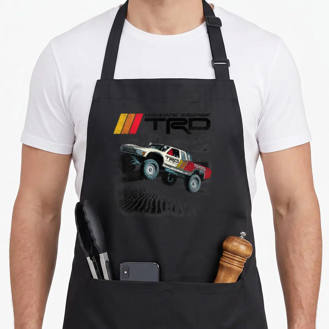 Iconic racing apron - Distressed TRD racing truck illustration with TOYOTA RACING DEVELOPMENT text in white red yellow — shown on a black cotton canvas apron against a white background