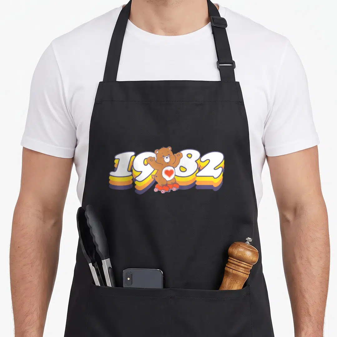 Funny bear apron - 1982 text with brown bear on roller skates in white, yellow, orange, and pink — shown on a black cotton canvas apron against a white background