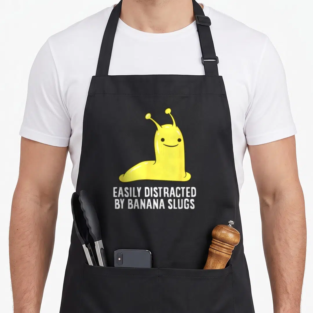 Slug banana apron - Cartoon yellow slug above white text reading EASILY DISTRACTED BY BANANA SLUGS — shown on a black cotton canvas apron against a white background