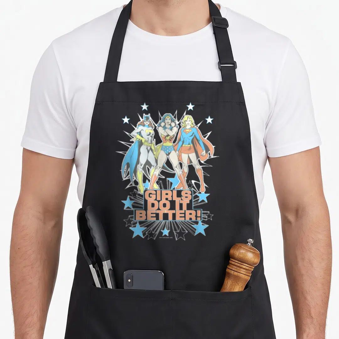 Supergirl batgirl apron - Three superheroines stand before a white burst above stacked text surrounded by blue stars — shown on a black cotton canvas apron against a white background