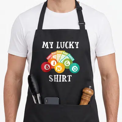 Funny lucky bingo apron - Five colorful bingo cards fanned behind balls spelling BINGO under MY LUCKY SHIRT text — shown on a black cotton canvas apron against a white background