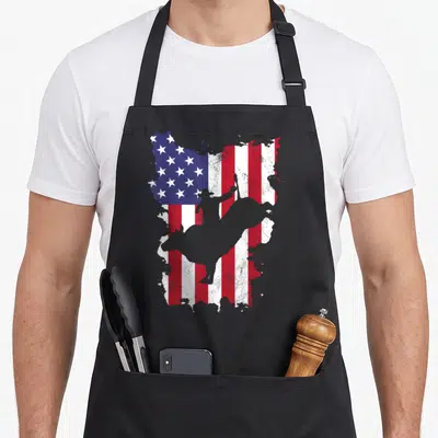 Bull 4th of july apron - Grey cowboy on bull silhouette over distressed red white and blue American flag pattern — shown on a black cotton canvas apron against a white background