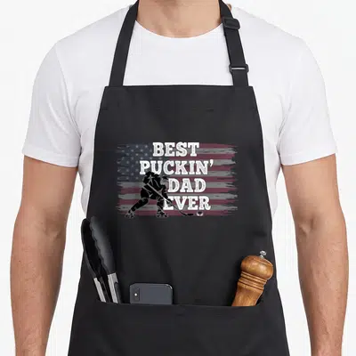 Puckin american flag apron - Distressed US flag with hockey player silhouette and best puckin dad ever text — shown on a black cotton canvas apron against a white background