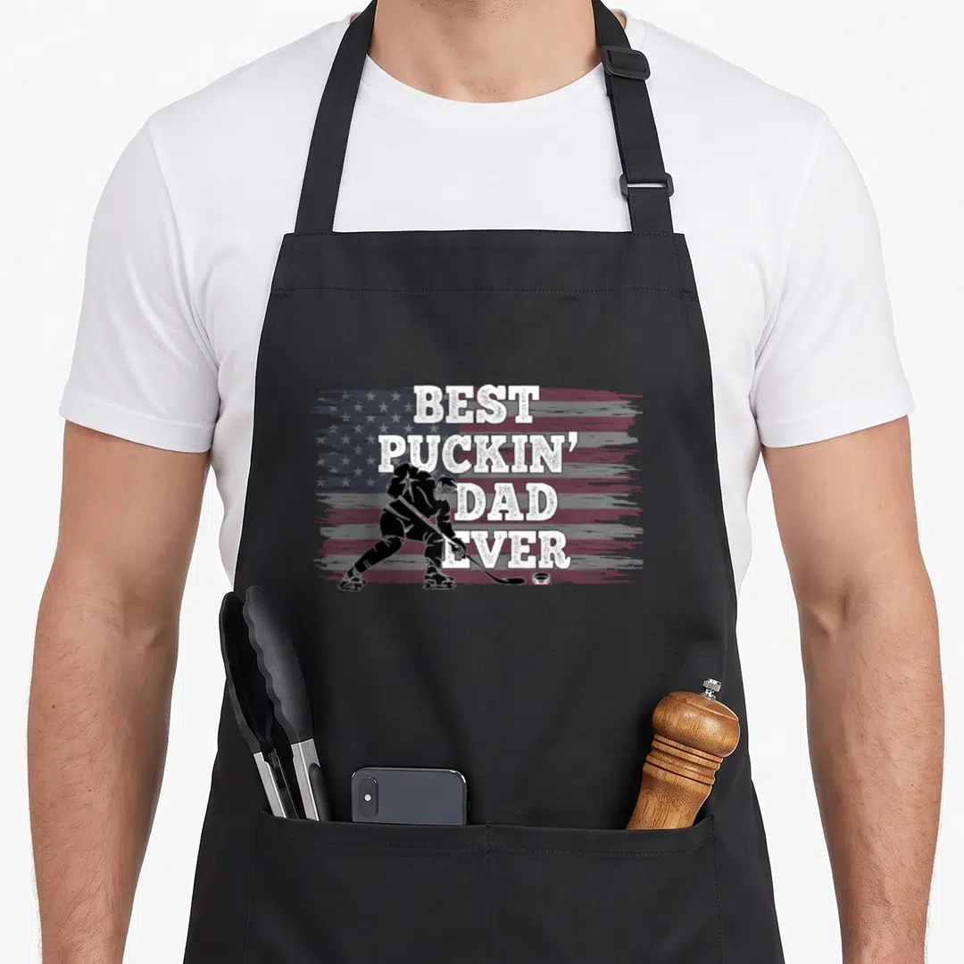 Puckin american flag apron - Distressed US flag with hockey player silhouette and best puckin dad ever text — shown on a black cotton canvas apron against a white background