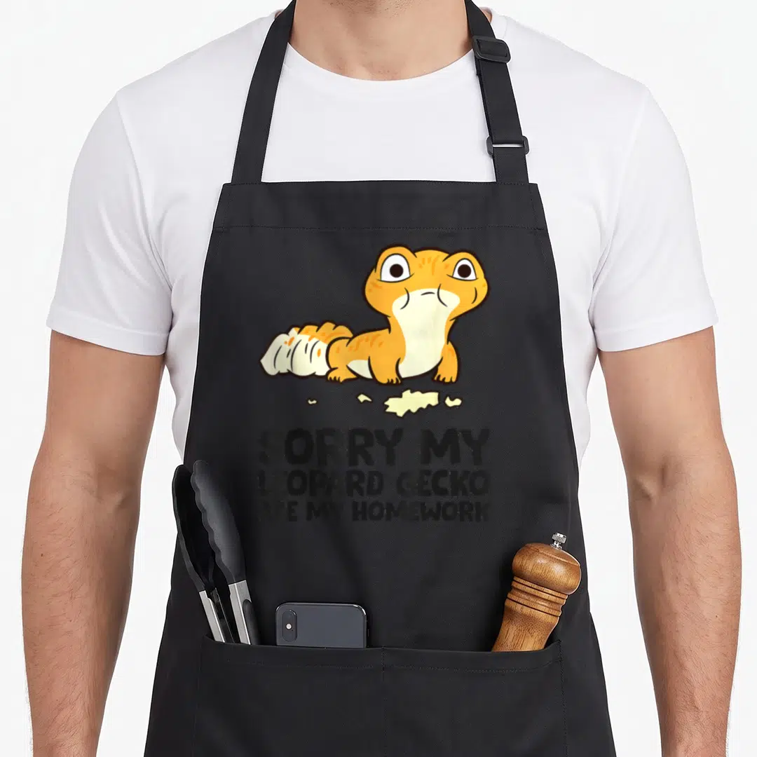 School leopard apron - Cartoon orange gecko above three paper scraps and black text apologizing for eaten homework — shown on a black cotton canvas apron against a white background