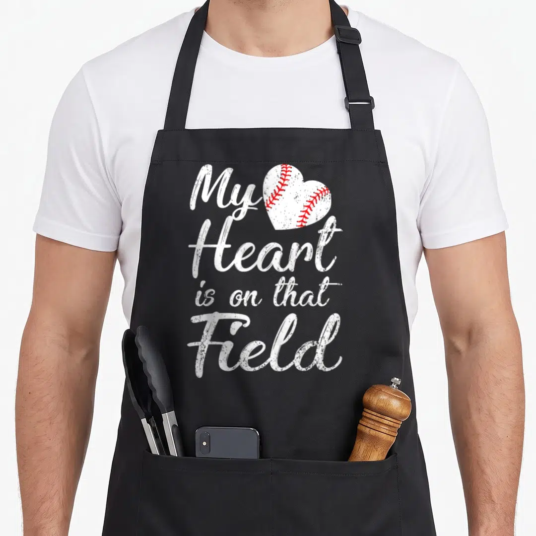 Field baseball apron - White script text My Heart is on that Field next to a red stitched heart baseball graphic — shown on a black cotton canvas apron against a white background