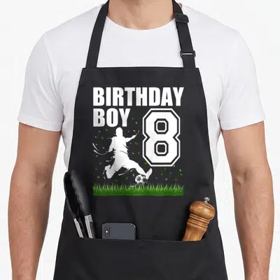 Theme 8th birthday apron - Soccer player silhouette kicking ball next to large number 8 and confetti on grass — shown on a black cotton canvas apron against a white background