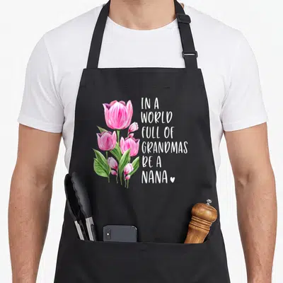 Nana tulip apron - Pink watercolor tulips beside stacked white text reading In A World Full Of Grandmas Be A Nana — shown on a black cotton canvas apron against a white background