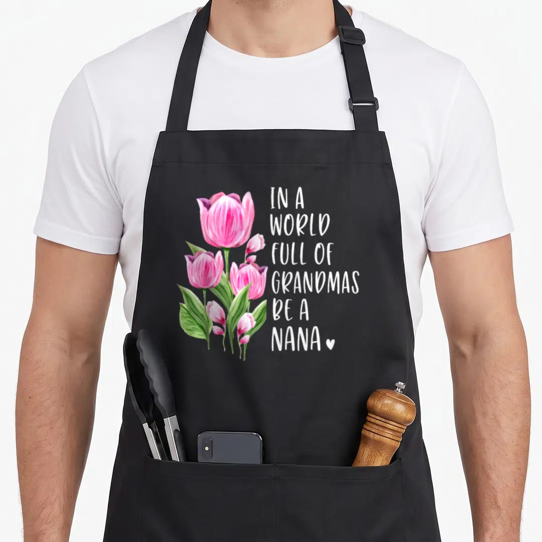 Nana tulip apron - Pink watercolor tulips beside stacked white text reading In A World Full Of Grandmas Be A Nana — shown on a black cotton canvas apron against a white background
