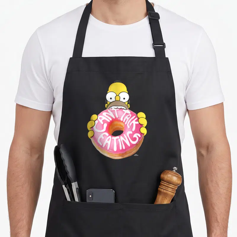 Homer donut apron - Cartoon head holding pink frosted donut with white text CAN'T TALK EATING — shown on a black cotton canvas apron against a white background