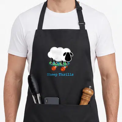 Thrill sheep apron - Cartoon sheep standing on skateboard with blue text Sheep Thrills below — shown on a black cotton canvas apron against a white background