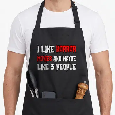 Movy horror apron - Distressed white and red text reading I like horror movies and maybe 3 people — shown on a black cotton canvas apron against a white background
