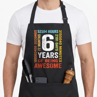 6th month birthday apron - Large white number 6 surrounded by colorful time measurements and years of being awesome text — shown on a black cotton canvas apron against a white background