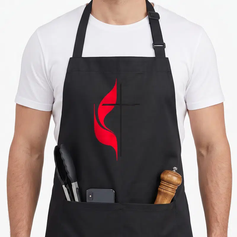 Methodist cross apron - Minimalist black cross with red flame shape curving around left side — shown on a black cotton canvas apron against a white background