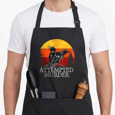 Attempted murder ravens apron - Two crows standing before a striped yellow orange and red sun above stacked grey — shown on a black cotton canvas apron against a white background
