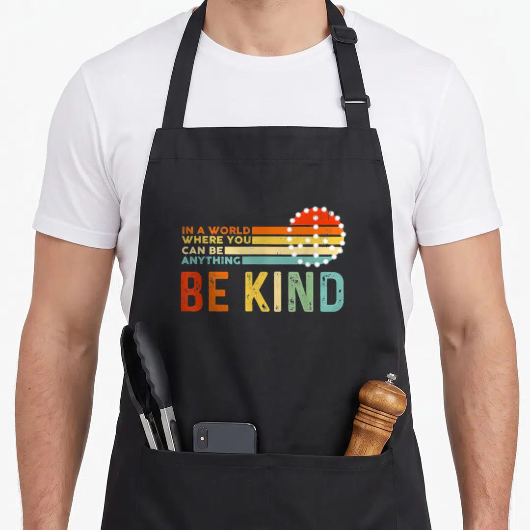 Kind hippie apron - Stacked Be Kind text with retro striped sun and white daisies in orange yellow teal — shown on a black cotton canvas apron against a white background