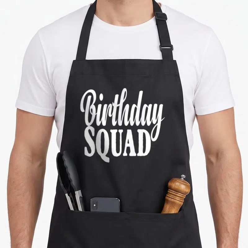 Funny squad birthday apron - White stacked text reading Birthday SQUAD in mixed fonts on grey background — shown on a black cotton canvas apron against a white background
