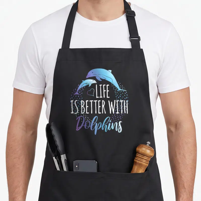 Better dolphin apron - Two blue and purple dolphins arching over a heart and text surrounded by water droplets — shown on a black cotton canvas apron against a white background