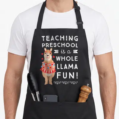 Appreciation preschool teacher apron - Cartoon llama with red blanket and text teaching preschool is a whole llama fun — shown on a black cotton canvas apron against a white background