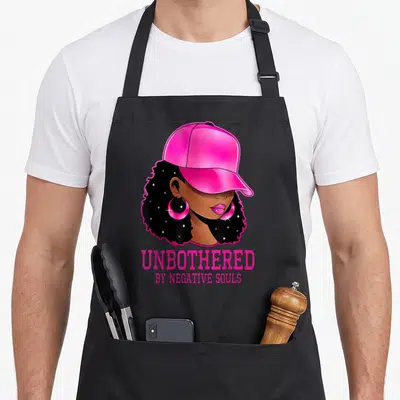 Sassy african american aprons - Cartoon woman in pink cap with UNBOTHERED BY NEGATIVE SOULS text — shown on a black cotton canvas apron against a white background