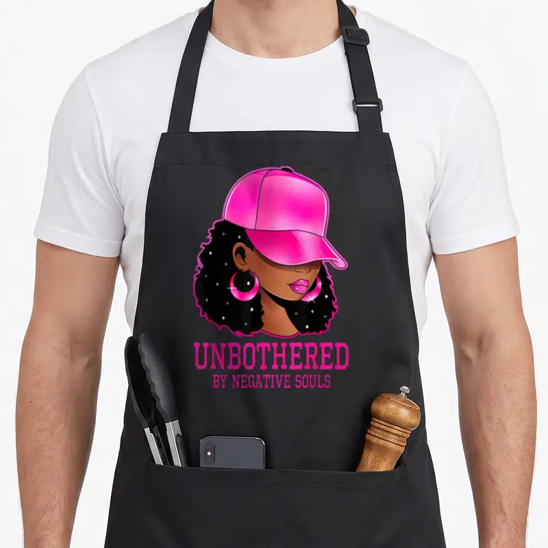 Sassy african american aprons - Cartoon woman in pink cap with UNBOTHERED BY NEGATIVE SOULS text — shown on a black cotton canvas apron against a white background