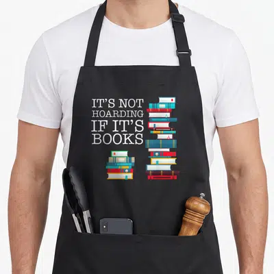 Hoarding sarcastic apron - White stacked text next to two piles of blue red and yellow books — shown on a black cotton canvas apron against a white background