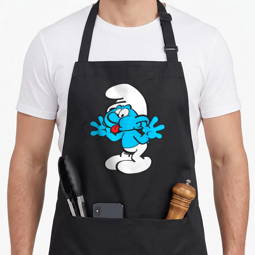 Jokey blue apron - Smurf character with tongue out, blue white red cartoon style — shown on a black cotton canvas apron against a white background