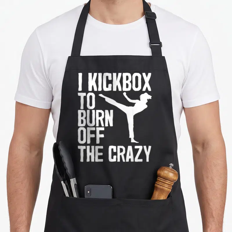 Kickboxing kick off apron - White stacked text I KICKBOX TO BURN OFF THE CRAZY beside a kicking fighter silhouette — shown on a black cotton canvas apron against a white background