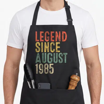 Legend august birthday apron - Stacked distressed text reading LEGEND SINCE AUGUST 1985 in orange yellow green and beige — shown on a black cotton canvas apron against a white background
