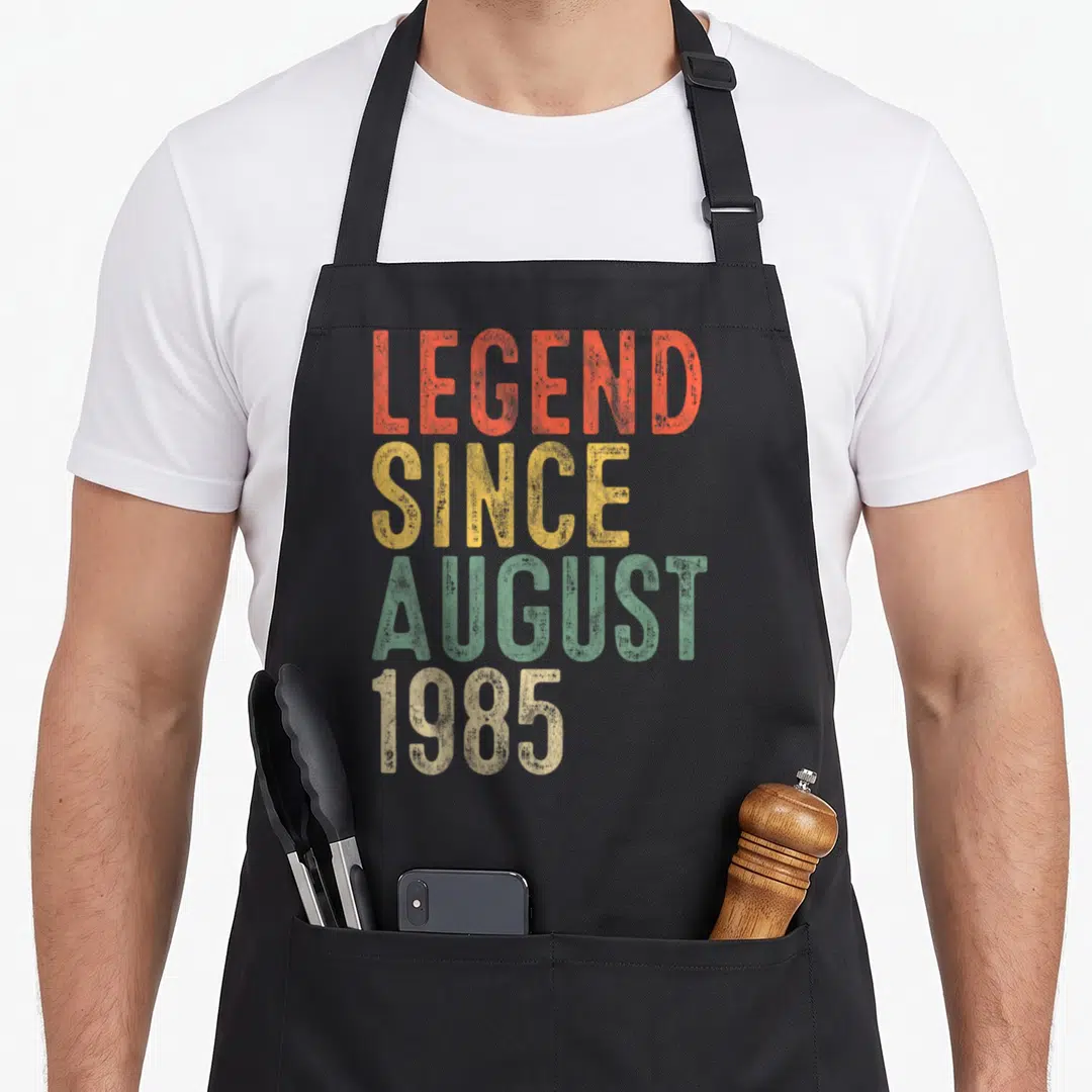 Legend august birthday apron - Stacked distressed text reading LEGEND SINCE AUGUST 1985 in orange yellow green and beige — shown on a black cotton canvas apron against a white background