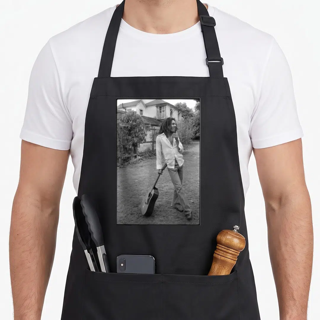 Bob david aprons - Grayscale photo of man holding guitar in yard with house background — shown on a black cotton canvas apron against a white background