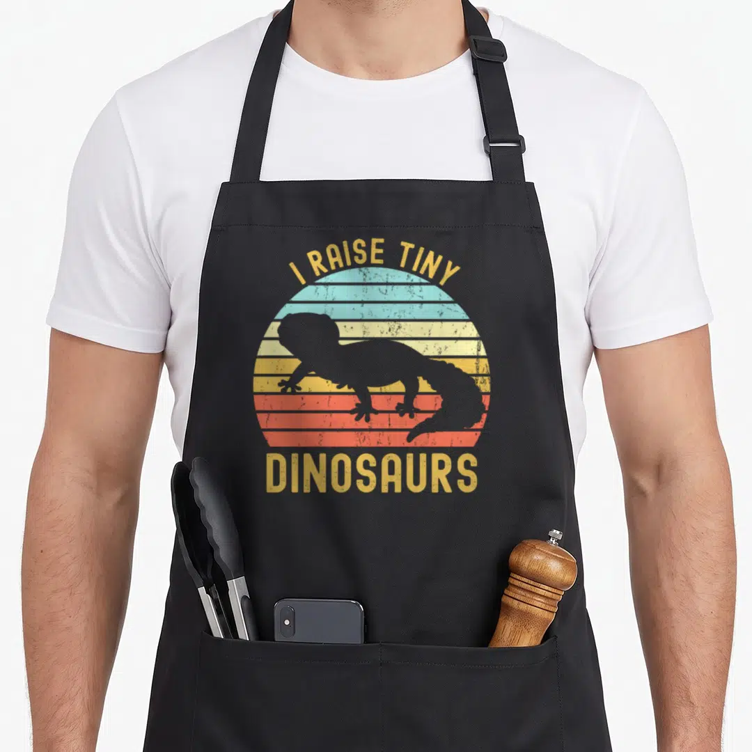 Tiny dinosaur leopard apron - Grey lizard silhouette over striped yellow orange red teal sun with arched text — shown on a black cotton canvas apron against a white background