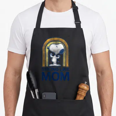 Strong mother's day aprons - Snoopy under yellow rainbow arch with STRONG LIKE MOM text in navy — shown on a black cotton canvas apron against a white background