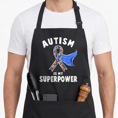 Superpower autism awareness apron - Cartoon ribbon with puzzle pieces and blue cape reading AUTISM IS MY SUPERPOWER — shown on a black cotton canvas apron against a white background