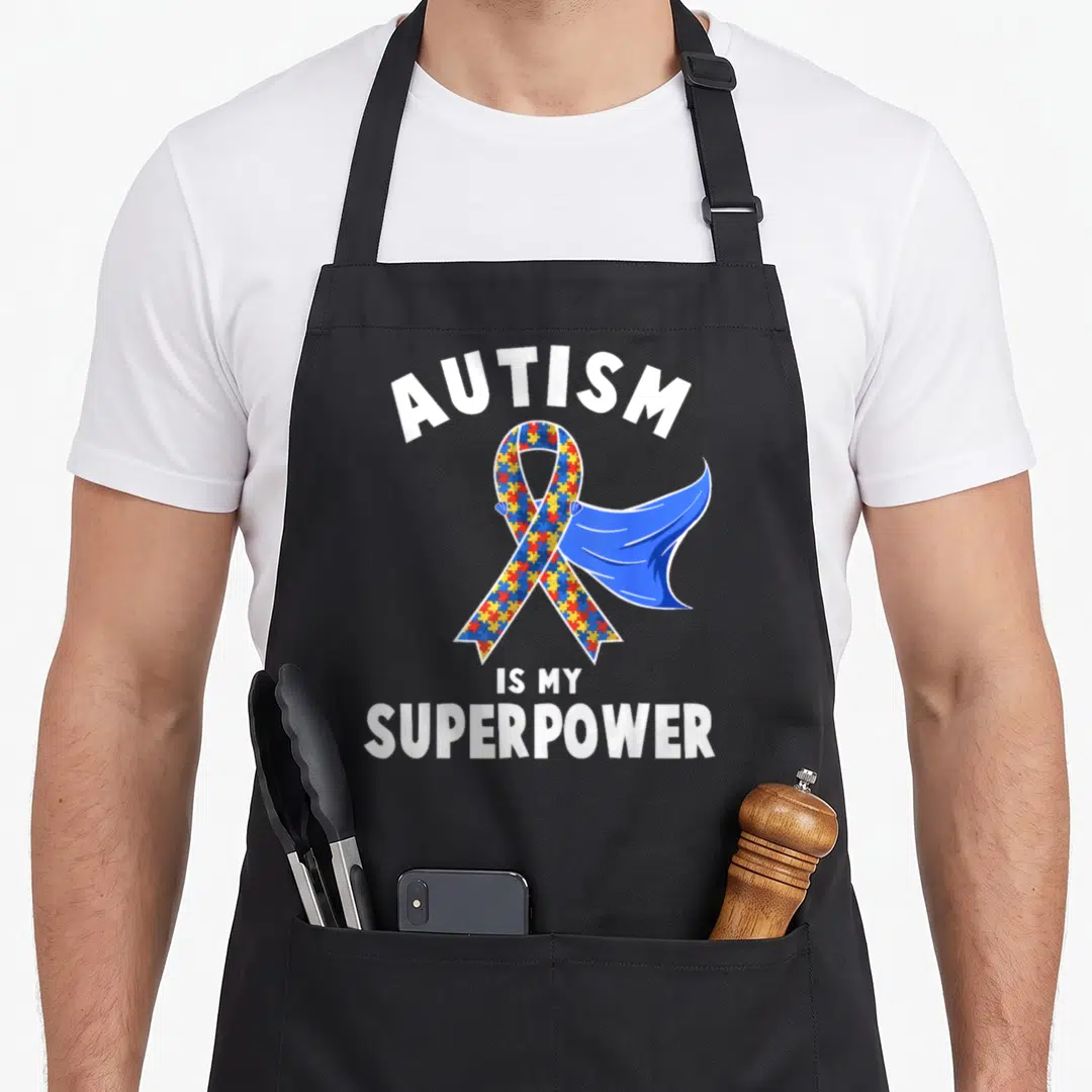 Superpower autism awareness apron - Cartoon ribbon with puzzle pieces and blue cape reading AUTISM IS MY SUPERPOWER — shown on a black cotton canvas apron against a white background