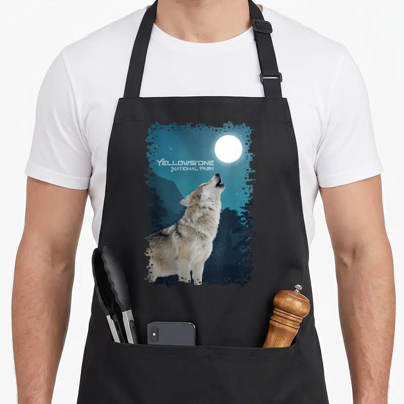 Souvenir national apron - Photo of a grey wolf howling at a white moon with stacked Yellowstone National Park text — shown on a black cotton canvas apron against a white background