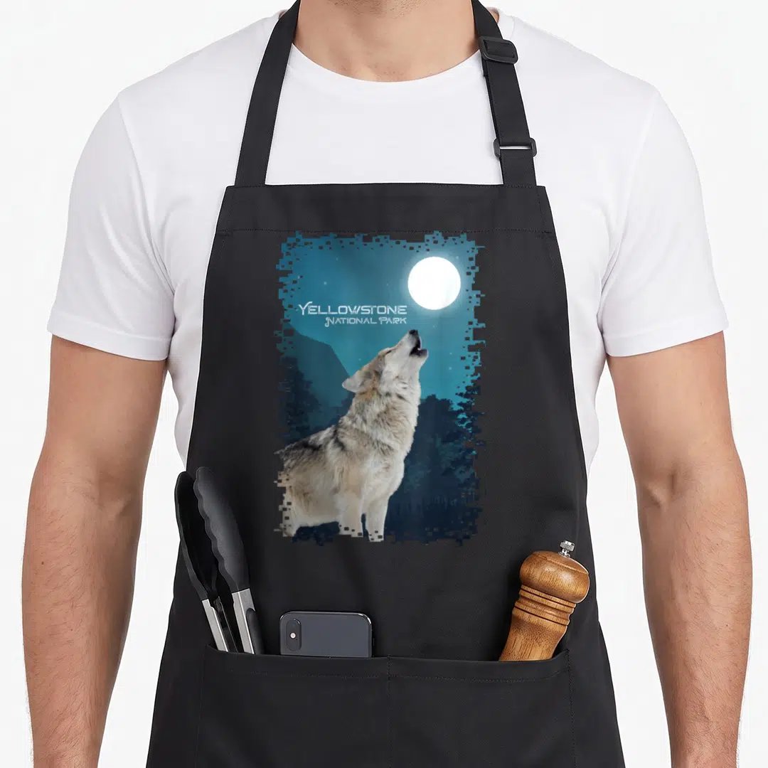 Souvenir national apron - Photo of a grey wolf howling at a white moon with stacked Yellowstone National Park text — shown on a black cotton canvas apron against a white background
